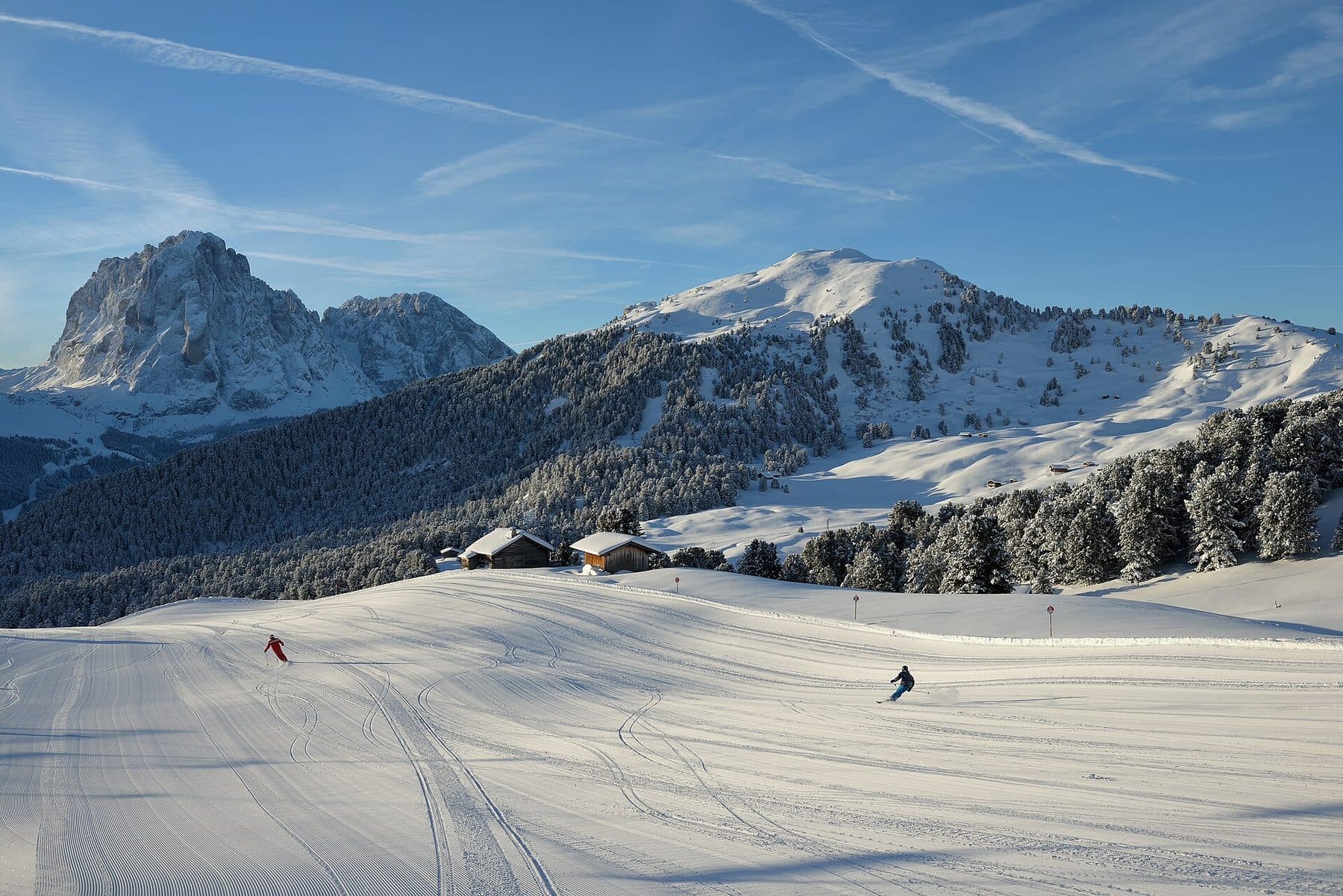 Wyratrakowana trasa w Val Gardena w Dolomitach: dwoje narciarzy, drewniane szałasy, ośnieżony las i skalisty masyw pod błękitnym niebem