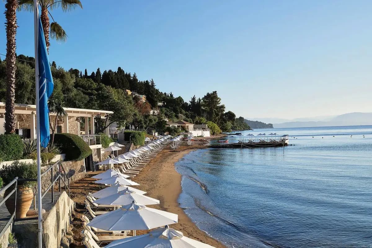 Plaża hotelu Aeolos Beach Resort w Peramie na Korfu: rząd leżaków pod białymi parasolami wzdłuż wąskiej, piaszczystej plaży nad spokojnym morzem