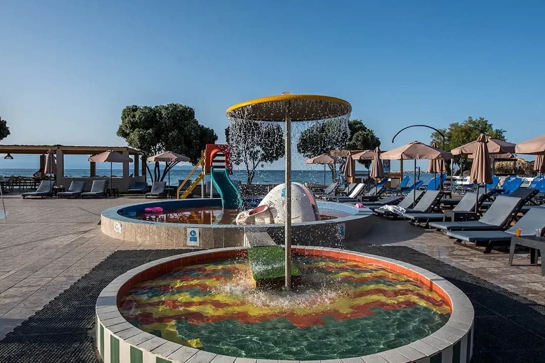 Brodzik z fontanną w kształcie parasola i mały basen ze zjeżdżalnią w hotelu Alykanas Beach w Grecji, w tle morze