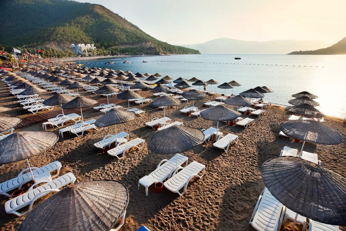 Plaża w İçmeler koło Marmaris z rzędami leżaków i parasoli, spokojna zatoka i porośnięte sosnami wzgórza w świetle zachodzącego słońca