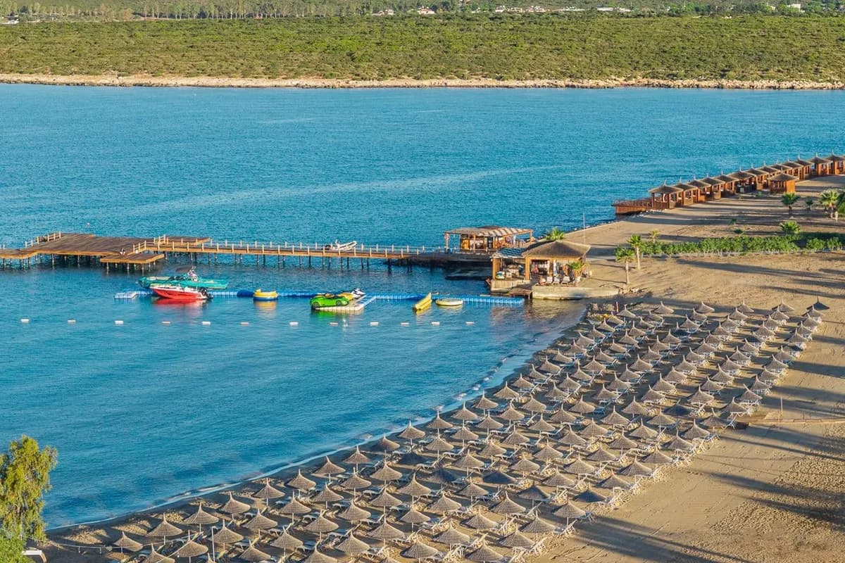 Hotelowa plaża w okolicy Didim: rzędy parasoli ze strzechy na piasku, drewniany pomost, łodzie i sprzęt wodny w zatoce, a za nią zielony brzeg