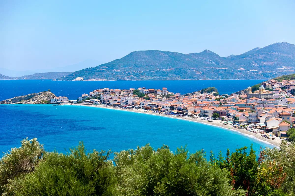 Widok z zalesionego wzgórza na Kokkari na Samos: miasteczko na cyplu, łuk kamienistej plaży i turkusowe morze, w tle góry