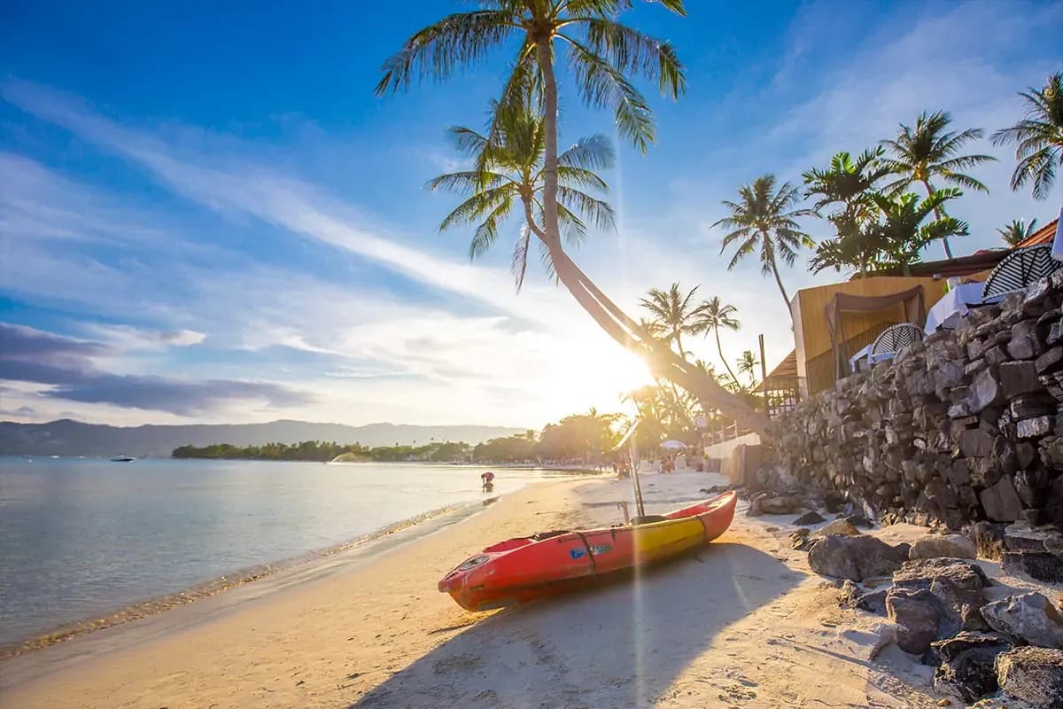 Piaszczysta plaża na Koh Samui w niskim słońcu: czerwono-żółty kajak na piasku, pochylona palma i kamienny mur hotelu nad spokojną zatoką