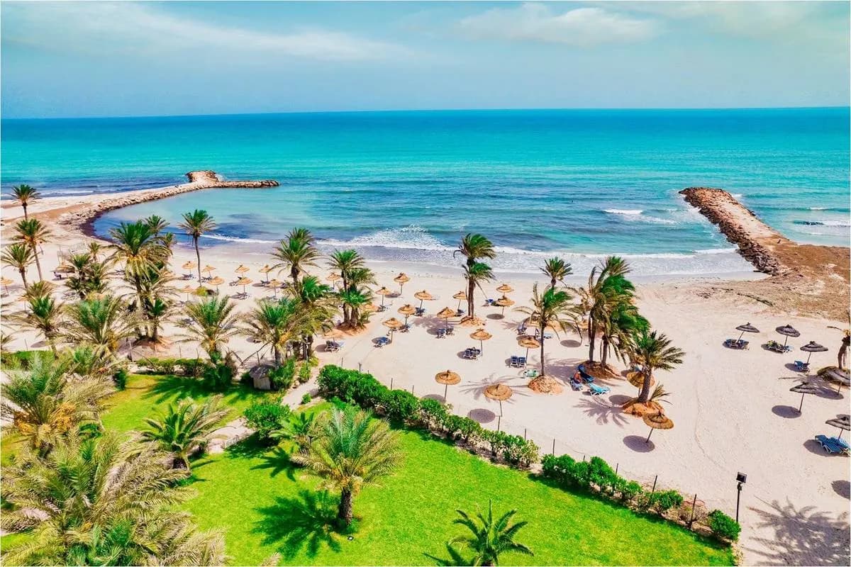 Piaszczysta plaża hotelu Club Palm Azur na Djerbie z palmami, parasolami z suchych liści i zatoką osłoniętą dwoma kamiennymi falochronami