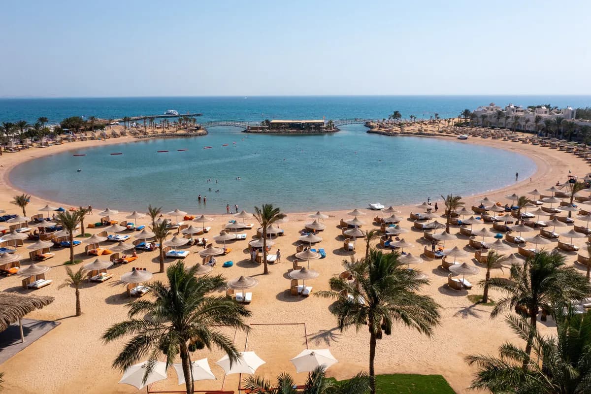 Półkolista zatoka przy hotelu Desert Rose w Hurghadzie: piaszczysta plaża z palmami i parasolami, pomosty prowadzące na wysepkę i Morze Czerwone