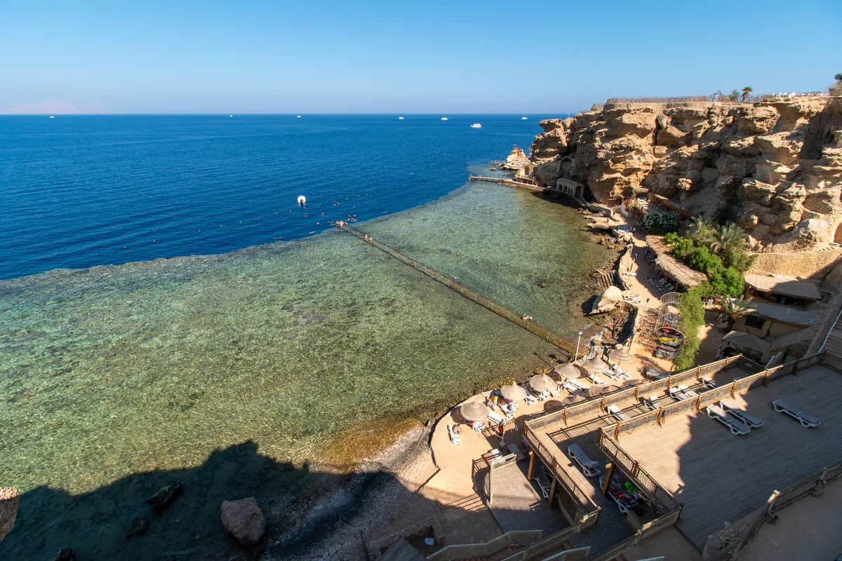 Rafa koralowa przy skalistym brzegu w Szarm el-Szejk: pomost prowadzi nad płytką rafą do głębokiej wody, obok plaża hotelu Dreams Beach z parasolami