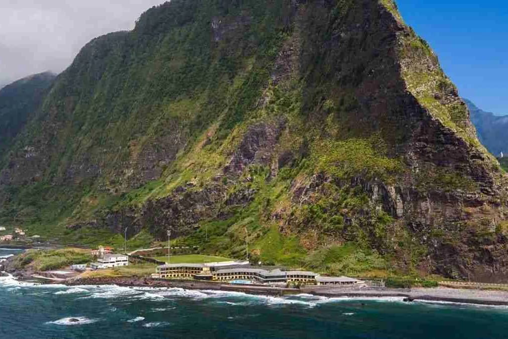 Strome, porośnięte zielenią zbocze nad oceanem w São Vicente na północnym wybrzeżu Madery, u jego podnóża hotel i kamienista plaża
