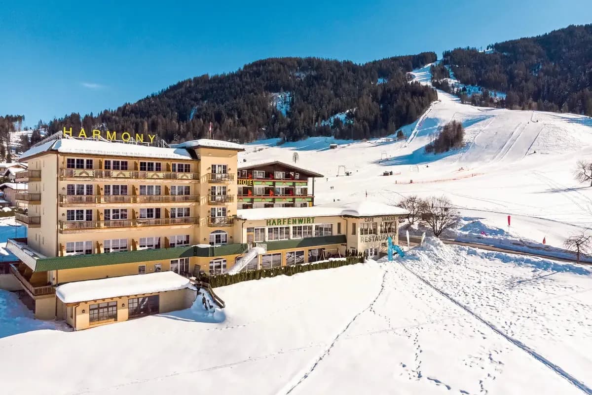 Hotel Harmony Harfenwirt w Niederau w Tyrolu zimą, u podnóża ośnieżonego stoku narciarskiego, w tle zalesione zbocza i błękitne niebo