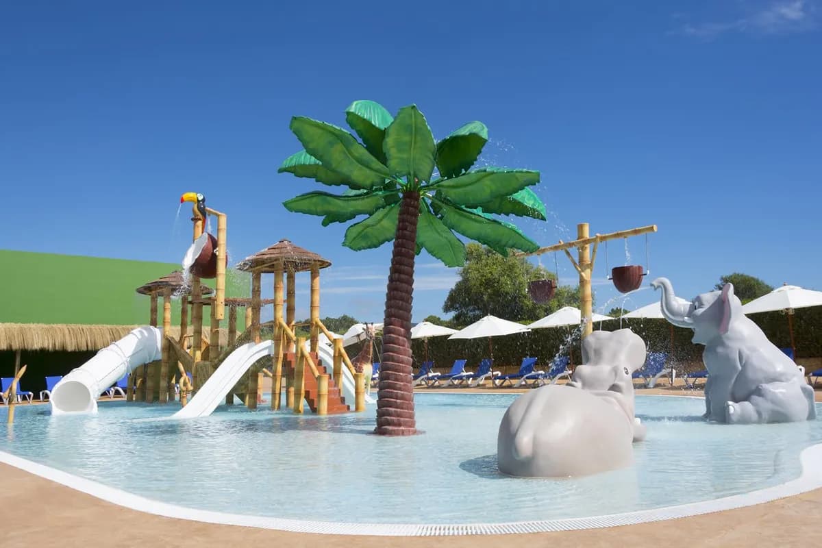 Brodzik dla dzieci w hotelu HSM Canarios Park na Majorce: zjeżdżalnie, sztuczna palma i figury słoni w płytkiej wodzie, w tle parasole i leżaki