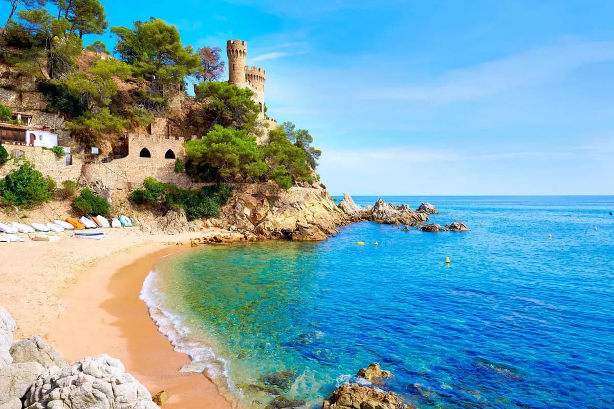 Plaża Sa Caleta w Lloret de Mar: złoty piasek, turkusowa woda i budowla w stylu zamku na skalistym, porośniętym sosnami cyplu