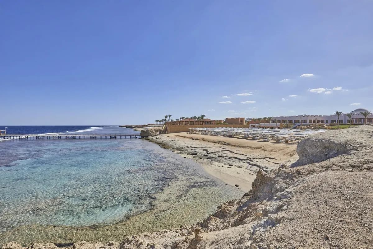 Płytka, przejrzysta woda nad rafą przy hotelu Jaz Neo Reef Marsa w Egipcie: skaliste wybrzeże, długi pomost w morzu i plaża z parasolami w oddali