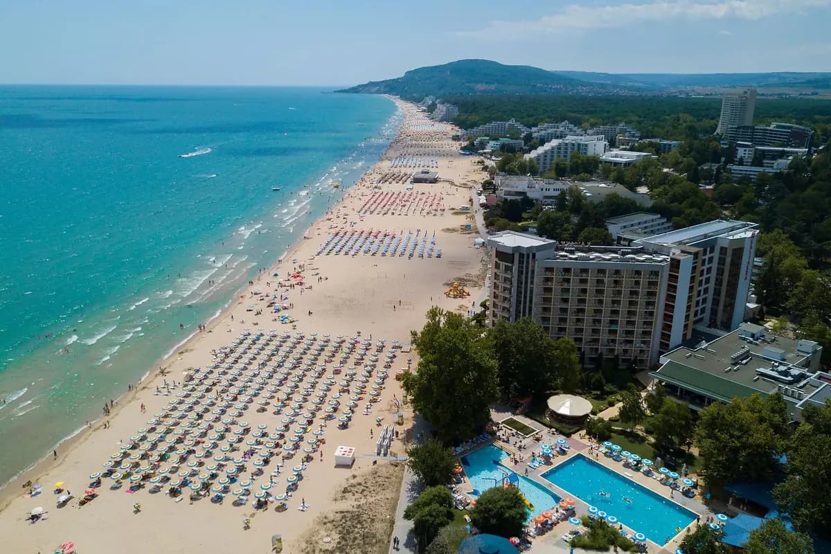 Szeroka, piaszczysta plaża w Albenie nad Morzem Czarnym z rzędami parasoli, hotelami i basenem, w tle zalesione wzgórza