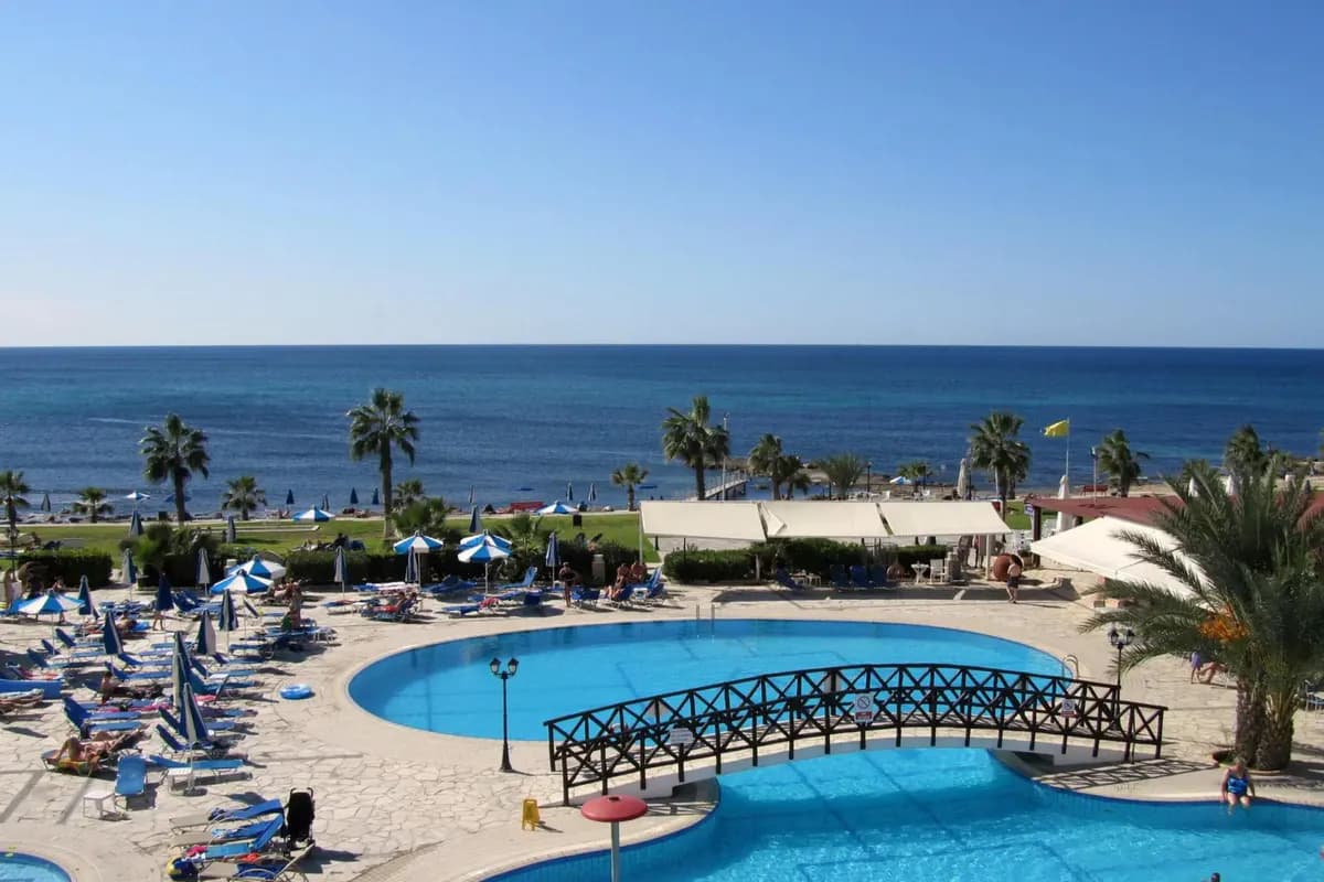 Basen z drewnianym mostkiem w hotelu Kefalos Beach w Pafos na Cyprze, za nim trawnik z palmami, plaża z parasolami i Morze Śródziemne