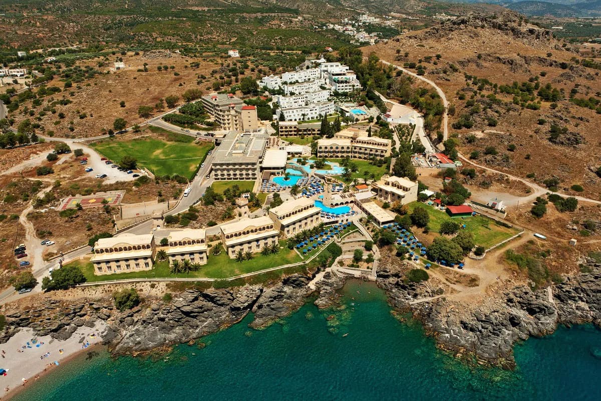 Hotel Lindos Royal na Rodos z lotu ptaka: budynki z basenami i niebieskimi parasolami nad skalistym brzegiem i turkusowym morzem, w tle suche wzgórza
