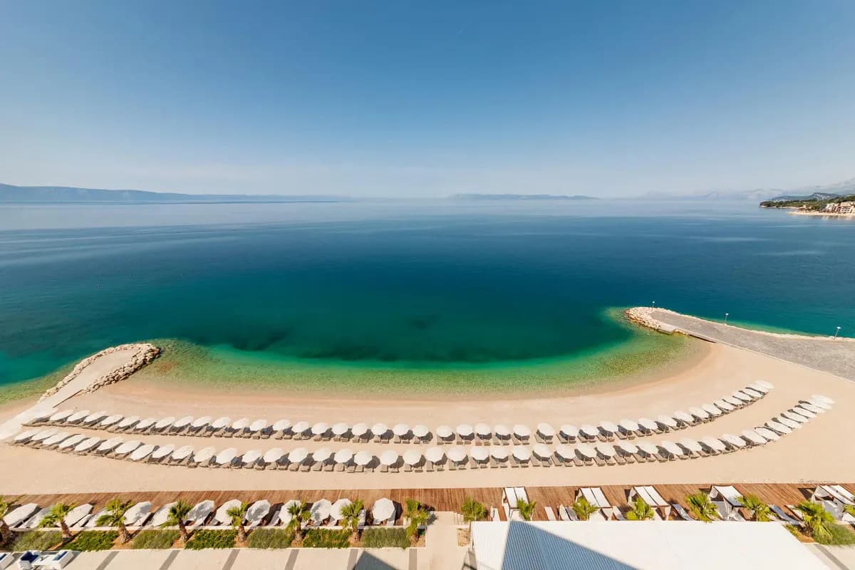 Żwirowa plaża z rzędami białych parasoli przy hotelu w Podgorze na Riwierze Makarskiej, przejrzyste morze i wyspy na horyzoncie