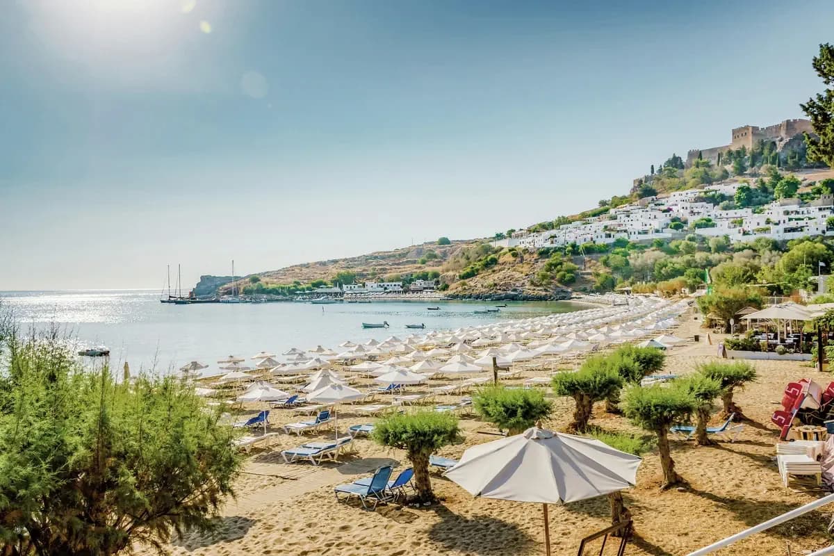 Piaszczysta plaża w zatoce Lindos na Rodos z parasolami i leżakami, w głębi białe domy miasteczka i mury akropolu na wzgórzu
