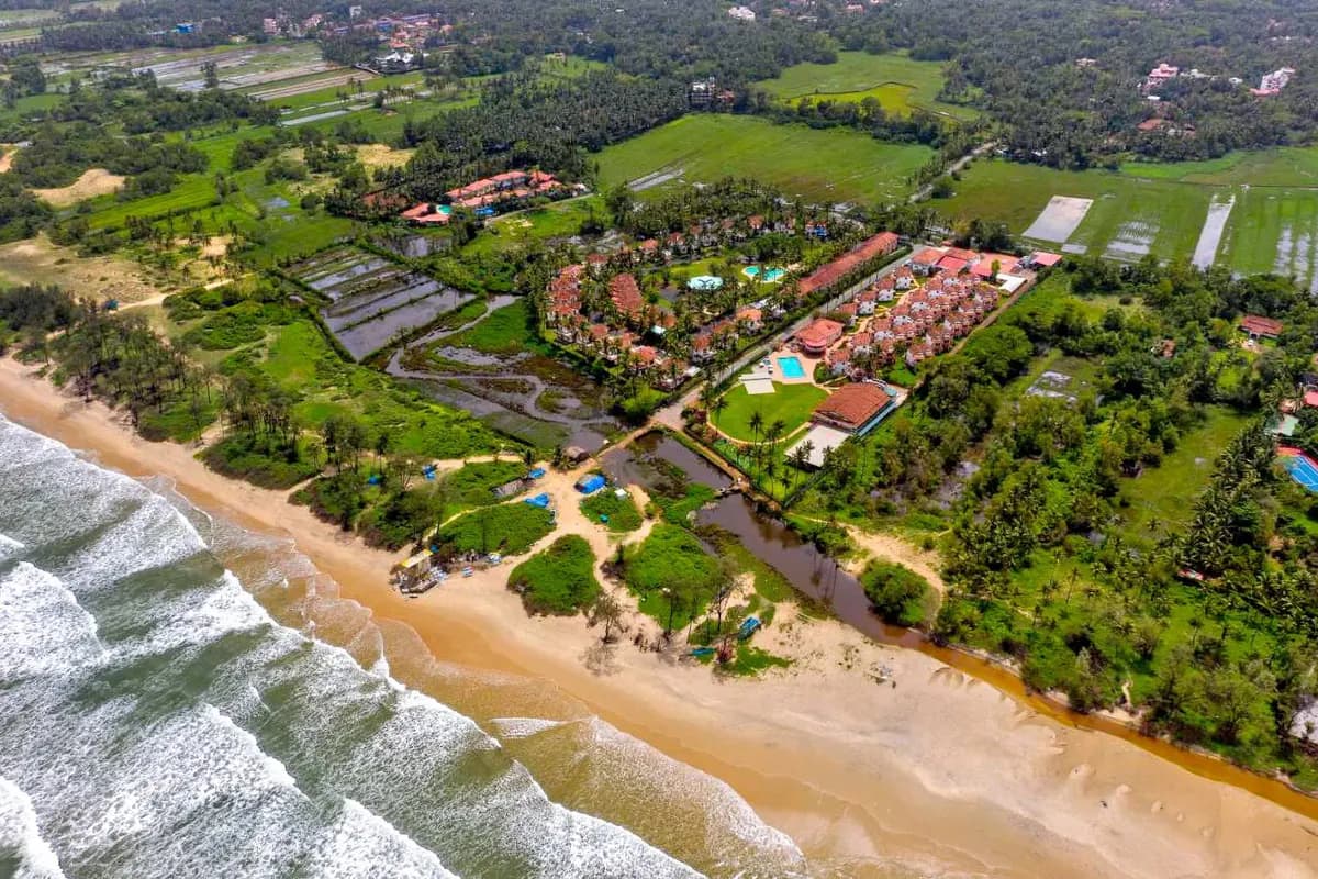 Widok z lotu ptaka na szeroką piaszczystą plażę w Goa z falami Morza Arabskiego, a za nią zabudowania hotelu Nanu Resort wśród palm i zielonych pól