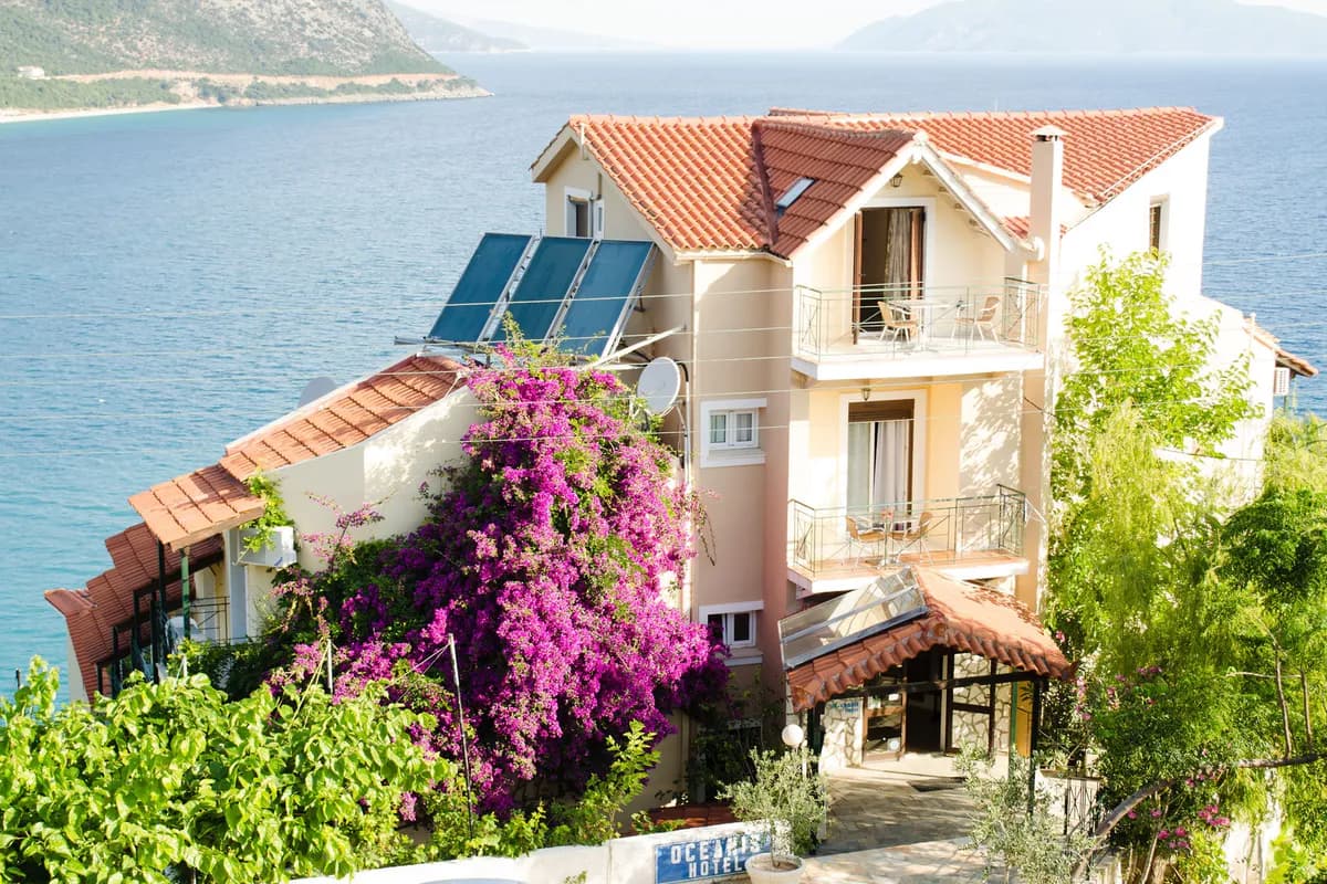 Hotel Oceanis w Poros na Kefalonii: budynek z czerwonym dachem i kwitnącą bugenwillą nad Morzem Jońskim, w tle górzyste wybrzeże
