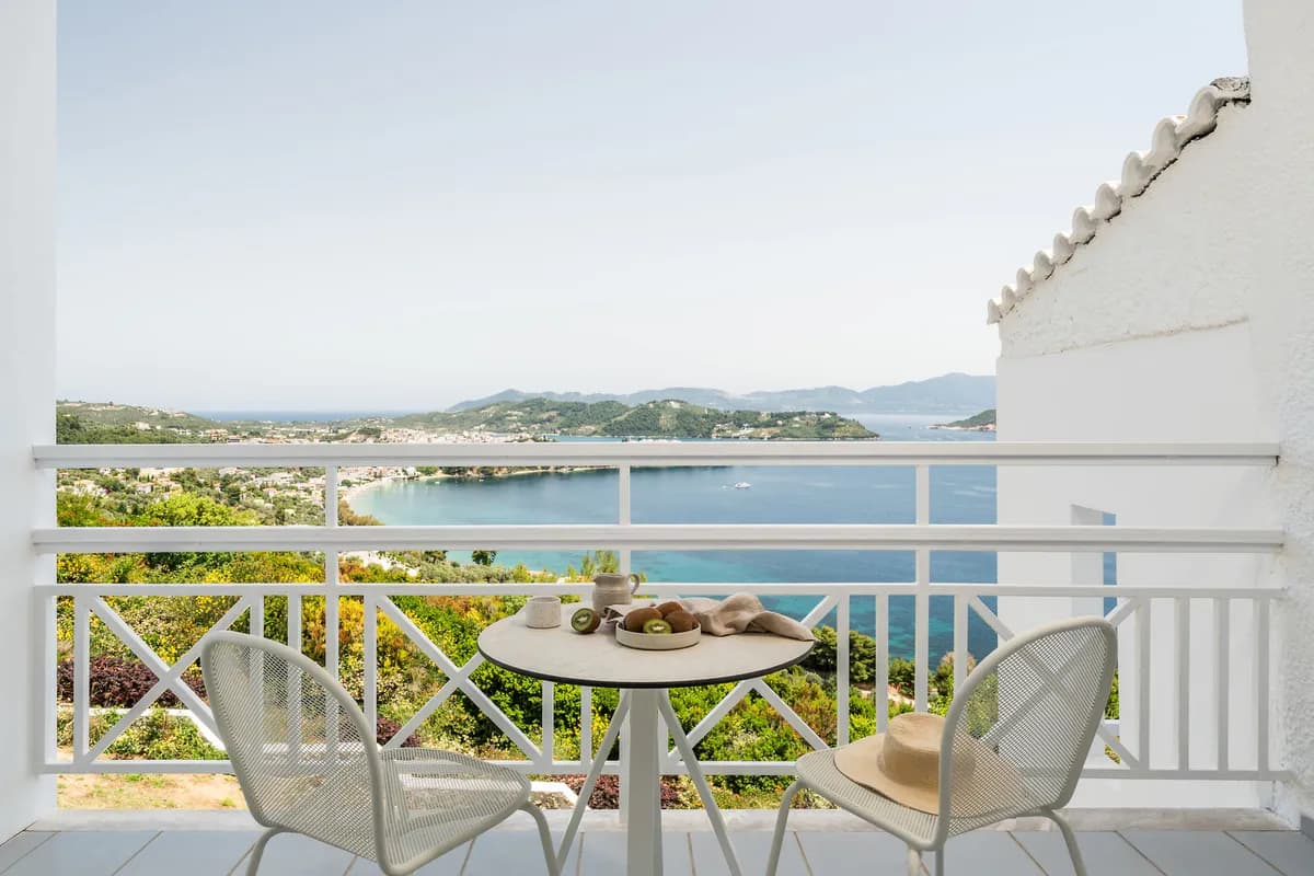 Balkon hotelu Oniro na Skiathos z widokiem na zatokę i zielone wzgórza: stolik z owocami kiwi i dzbankiem, słomkowy kapelusz na krześle
