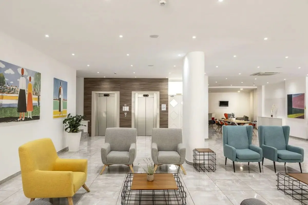 Lobby hotelu Papantonia na Cyprze: kolorowe fotele przy niskich stolikach, dwie windy i obrazy na białych ścianach
