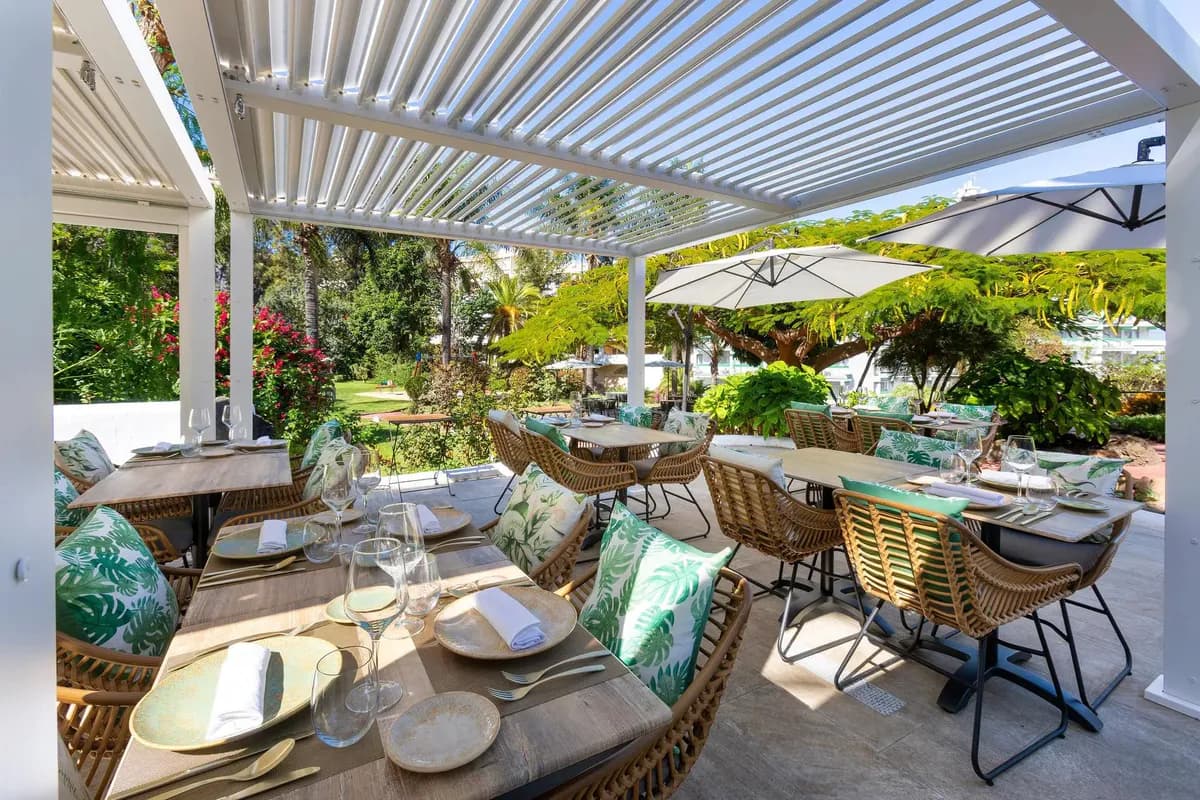 Nakryte stoliki na tarasie restauracji hotelu Parque Vacacional Eden na Teneryfie, pod białą pergolą, wśród palm i kwitnących krzewów