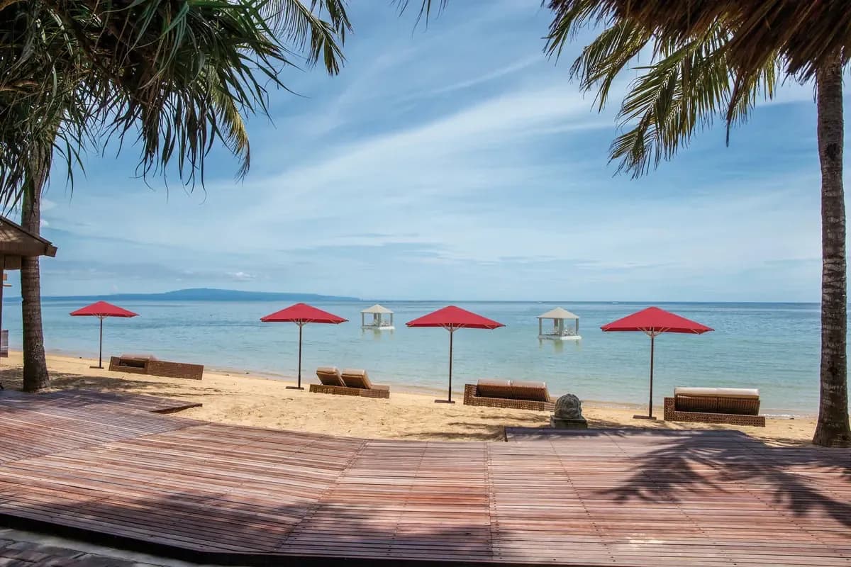 Plaża hotelu Puri Santrian w Sanur na Bali z czerwonymi parasolami, leżakami, palmami i drewnianym tarasem, w tle spokojne morze