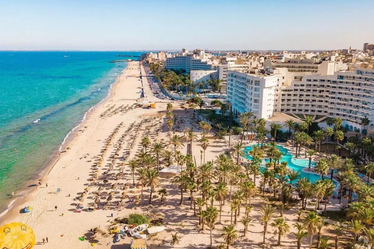 Hotel Riadh Palms w Sousse z lotu ptaka: długa piaszczysta plaża z parasolami, ogród palmowy z basenem i rząd hoteli nad turkusowym morzem