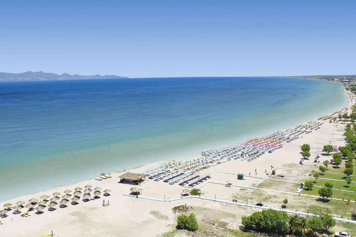 Długa, piaszczysta plaża w Tigaki na Kos z lotu ptaka: jasna, płytka woda przy brzegu, rzędy parasoli i góry na horyzoncie