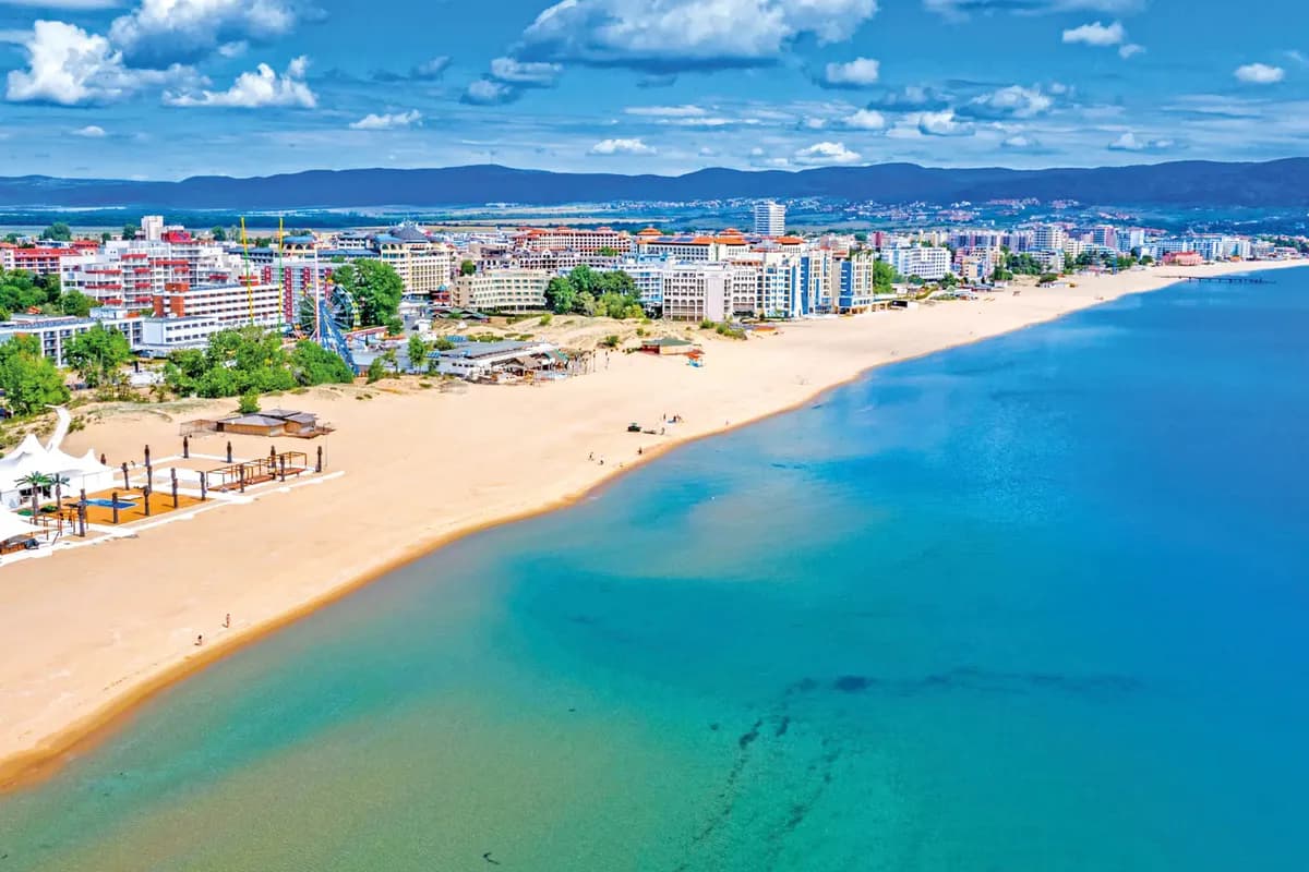 Słoneczny Brzeg z lotu ptaka: szeroka piaszczysta plaża, turkusowa płycizna, hotele i diabelski młyn, w tle pasmo gór