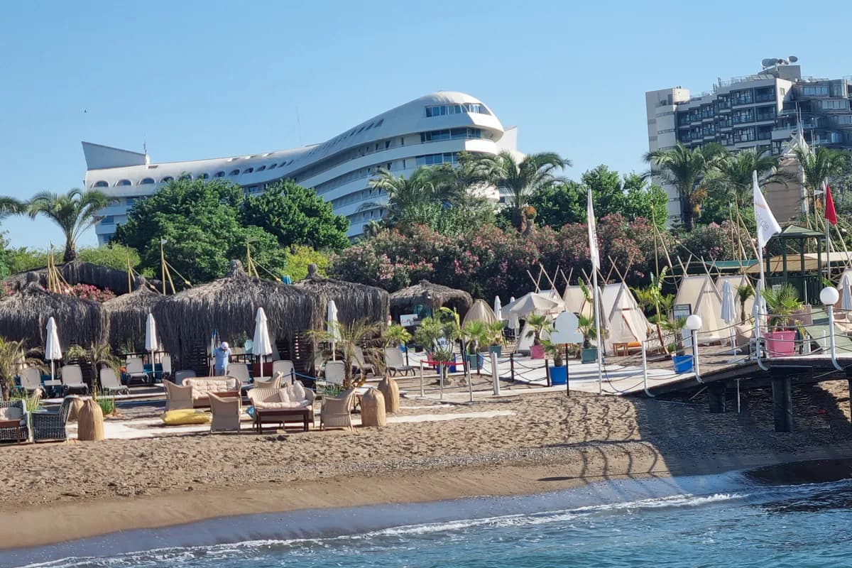 Biały budynek hotelu Concorde De Luxe Resort w Larze w kształcie samolotu Concorde, widziany z plaży z altanami krytymi strzechą