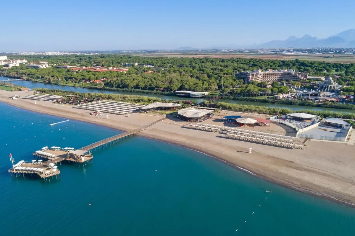 Plaża w Belek z lotu ptaka: szeroki piaszczysty brzeg z pomostem, za nim rzeka i sosnowy las z hotelami, w oddali góry Taurus