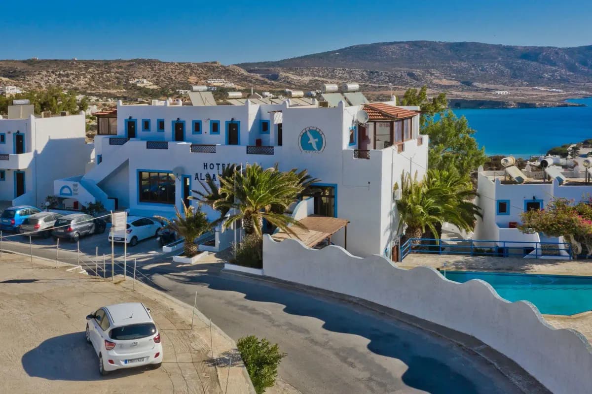 Biały hotel Albatros w Amoopi na Karpathos z niebieskimi okiennicami i palmami, w tle turkusowa zatoka i suche, kamieniste wzgórza