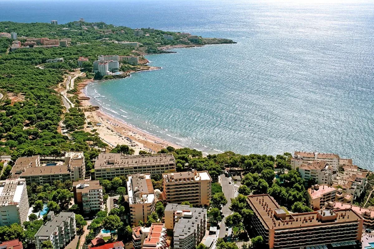 Salou z lotu ptaka: hotele wśród sosen, piaszczysta plaża w łukowatej zatoce, spokojne morze i skalisty cypel w oddali