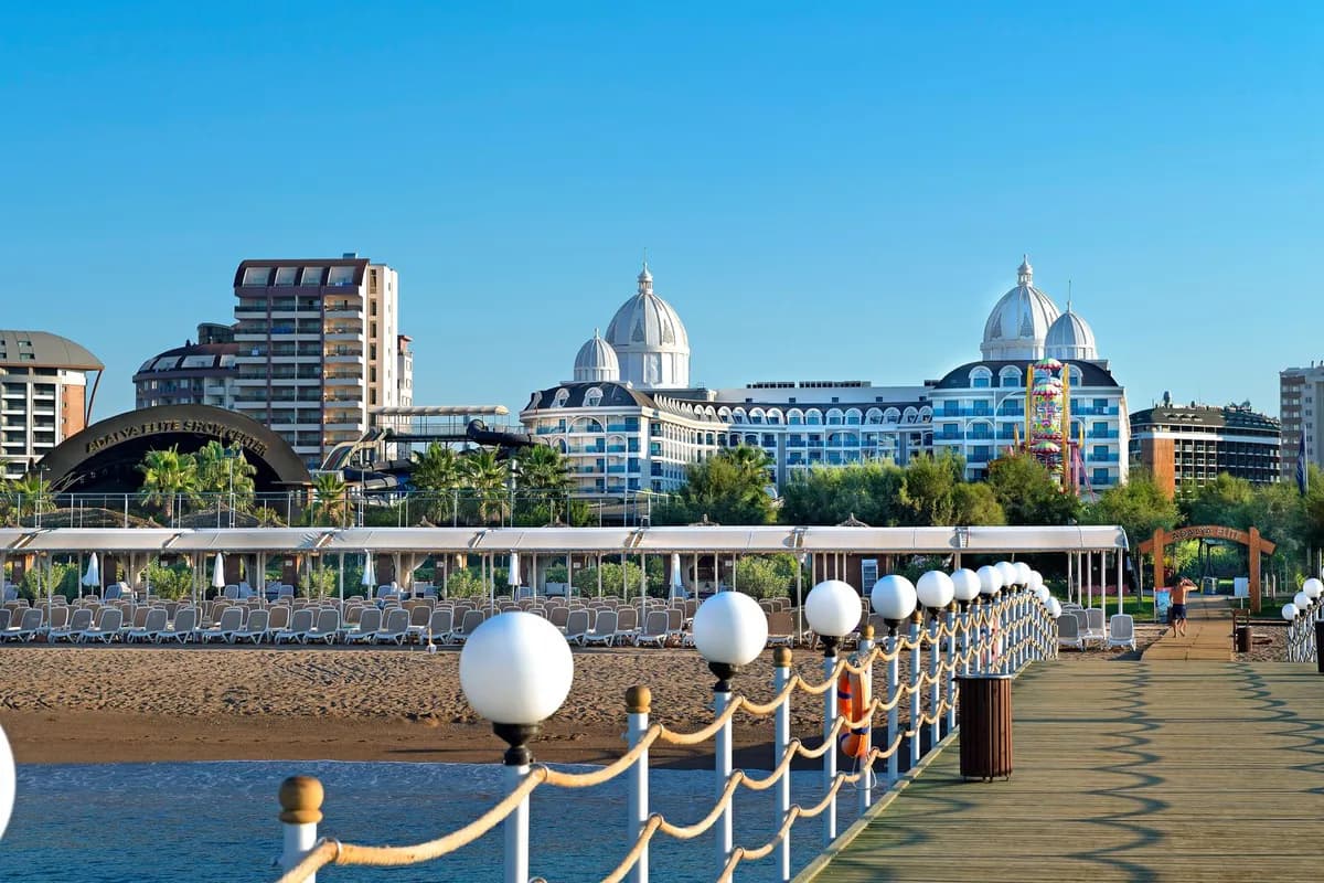 Widok z drewnianego pomostu na piaszczystą plażę z leżakami pod zadaszeniem i hotel Adalya Elite Lara z białymi kopułami w Larze pod Antalyą