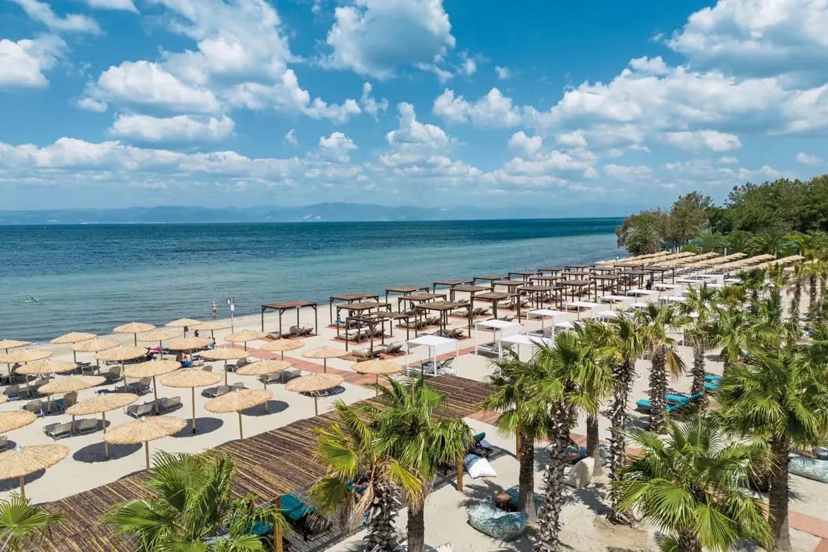 Piaszczysta plaża w Skala Prinos na Thassos z parasolami ze strzechy i palmami, w oddali góry na wybrzeżu Grecji kontynentalnej