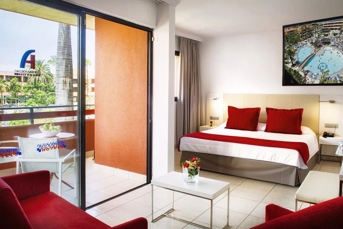 Pokój hotelu Alexandre La Siesta na Teneryfie: podwójne łóżko z czerwonymi poduszkami, kanapa, stolik i otwarte drzwi na balkon z widokiem na palmy