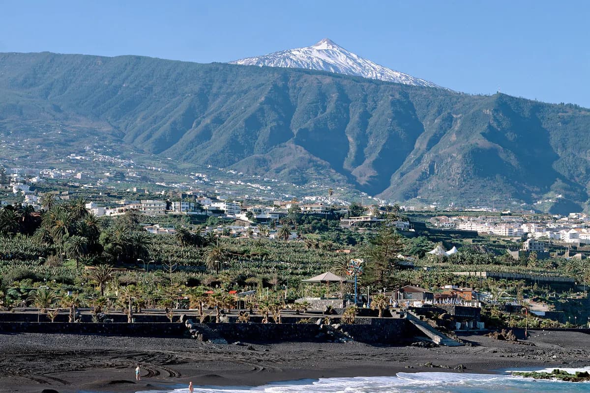 Plaża z ciemnym wulkanicznym piaskiem w Puerto de la Cruz na Teneryfie, za nią palmy, zabudowa doliny i ośnieżony szczyt Teide