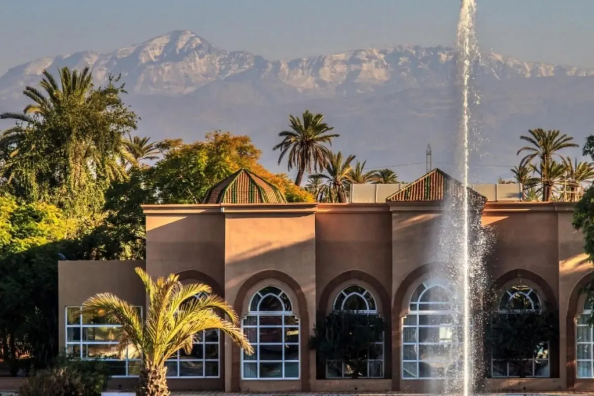 Budynek hotelu Barceló Palmeraie Oasis Resort w Marrakeszu z łukowymi oknami, fontanną i palmami, w tle ośnieżone szczyty Atlasu