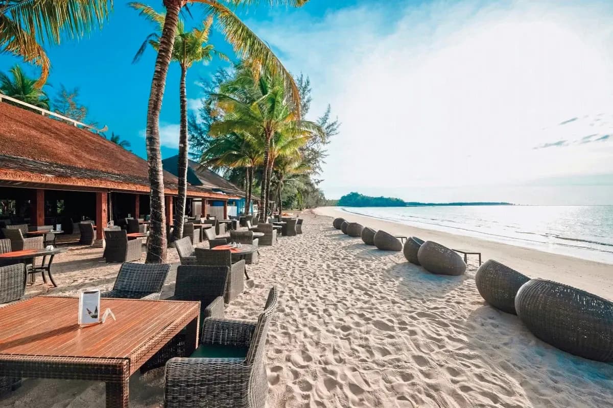 Szeroka piaszczysta plaża przy hotelu Beyond Resort Khao Lak: wiklinowe fotele i stoliki pod palmami, spokojne morze i zalesiony cypel w oddali
