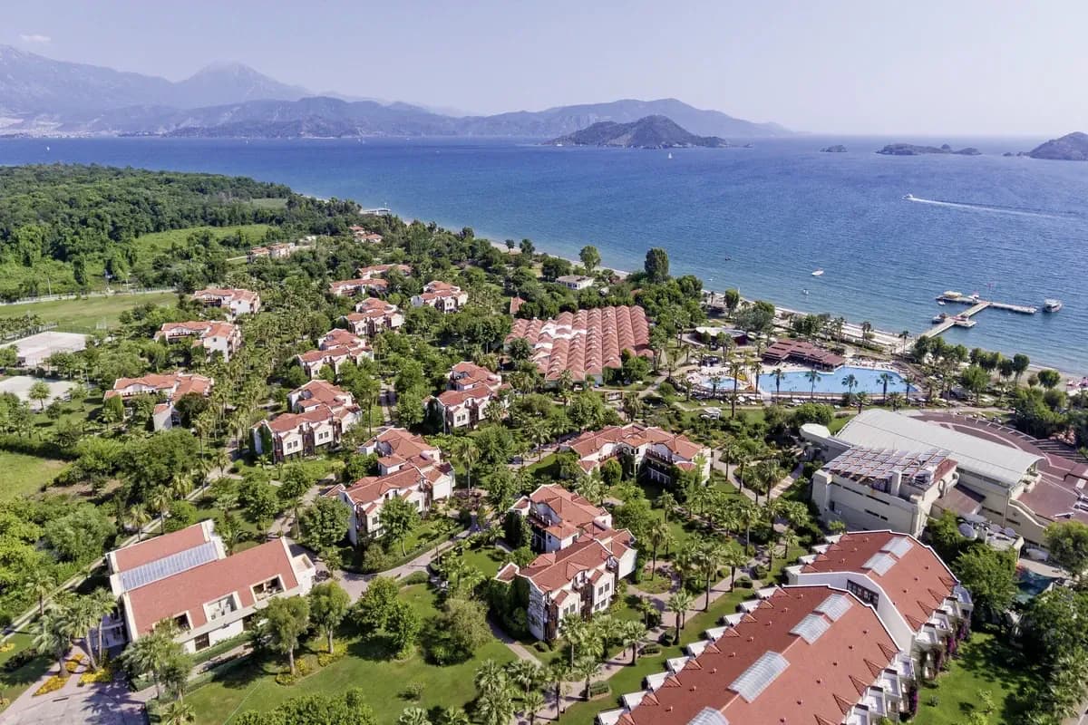Widok z lotu ptaka na hotel wśród palm nad Zatoką Fethiye: piaszczysta plaża z pomostem, granatowe morze, wysepki i góry na horyzoncie