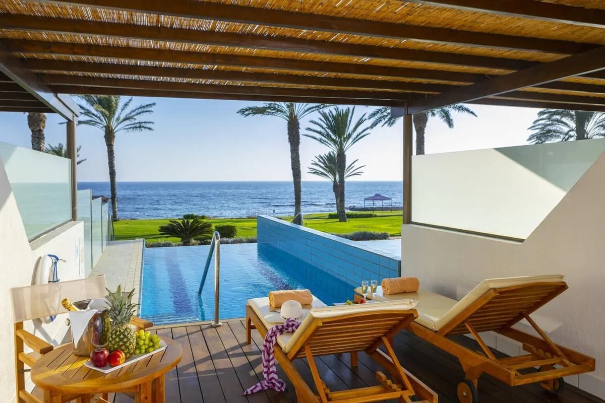 Zadaszony taras pokoju w hotelu Constantinou Bros Athena Beach w Pafos: dwa drewniane leżaki, prywatny basen, palmy na trawniku i morze w tle