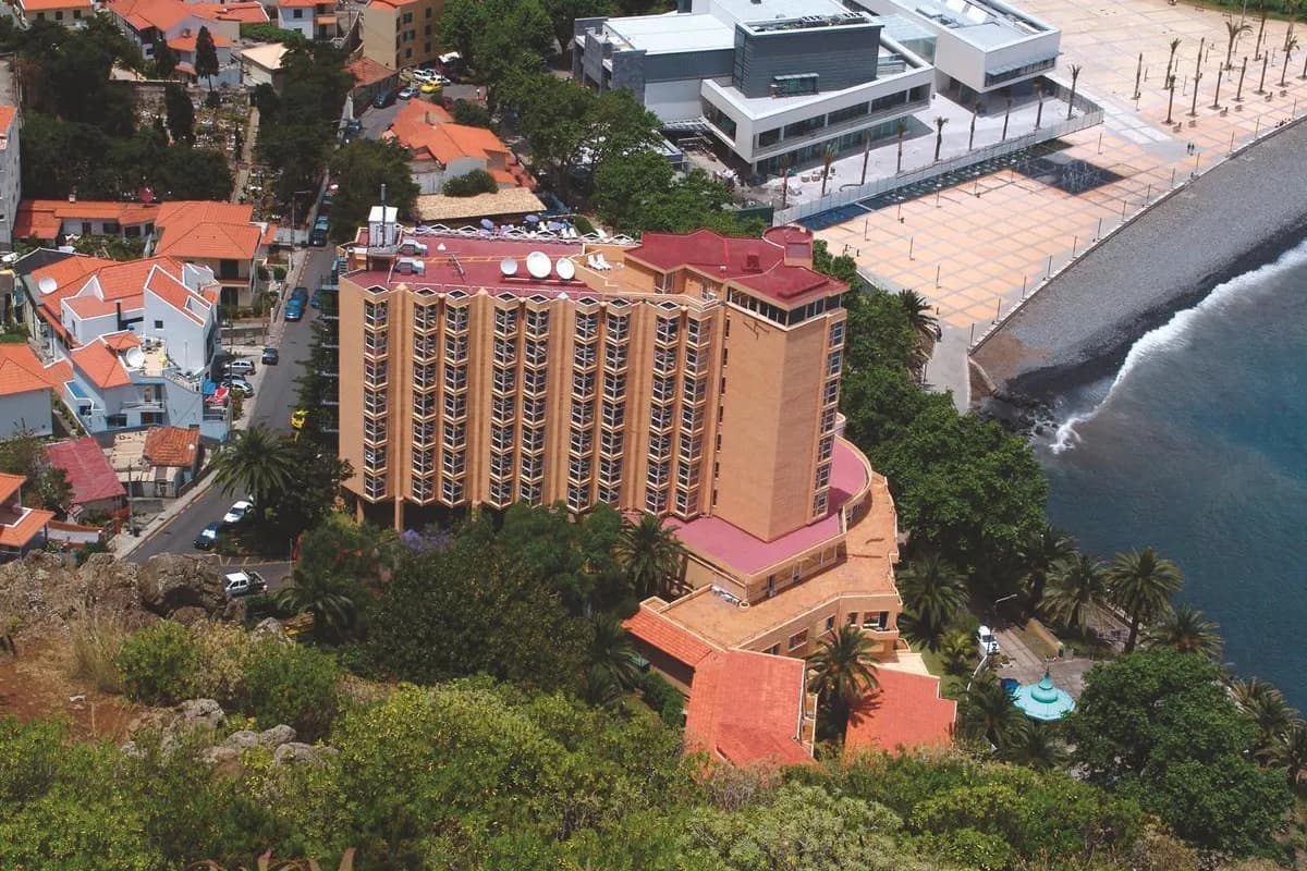 Hotel Dom Pedro Madeira na Maderze z lotu ptaka: wysoki budynek wśród palm i domów z czerwonymi dachami, obok nadmorski plac i kamienisty brzeg oceanu
