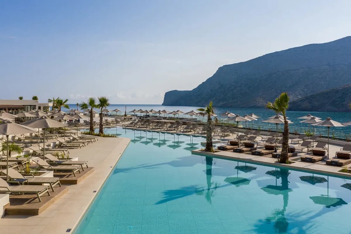 Długi basen z leżakami, parasolami i palmami nad morzem w hotelu Fodele Beach na Krecie, w tle skaliste wzgórza schodzące do zatoki
