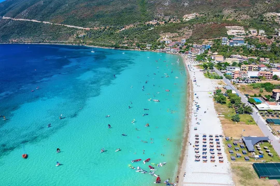 Zatoka Vasiliki na Lefkadzie z lotu ptaka: długa, jasna plaża z parasolami, turkusowa woda z deskami windsurfingowymi i zabudowa na zboczu góry