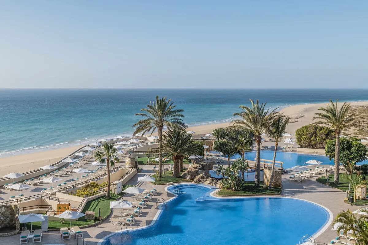 Baseny i palmy hotelu Iberostar Waves Playa Gaviotas na Fuerteventurze, a za nimi długa piaszczysta plaża i ocean