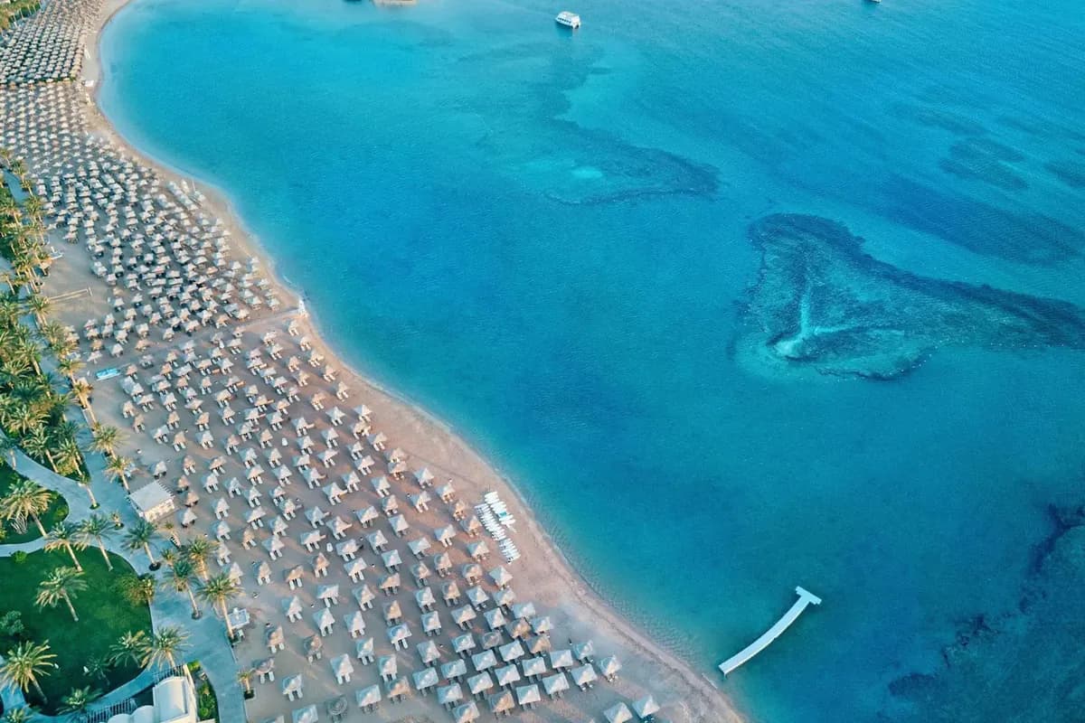 Plaża hotelu Jaz Makadi Saraya w Makadi Bay z lotu ptaka: rzędy parasoli, palmy, pomost i ciemne plamy rafy w turkusowej wodzie