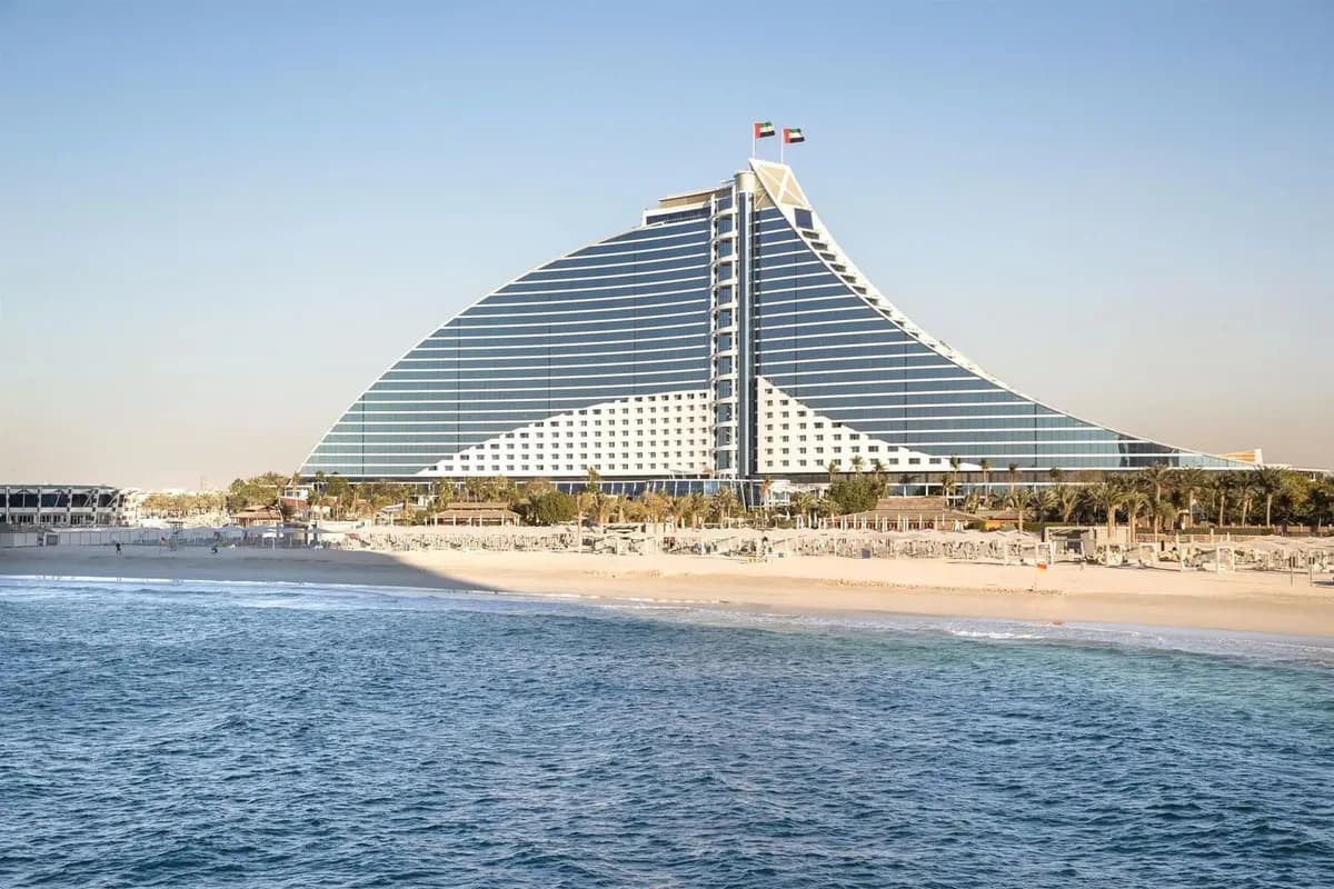Hotel Jumeirah Beach w Dubaju o kształcie fali, przed nim szeroka, piaszczysta plaża z parasolami i palmami, na pierwszym planie spokojne morze