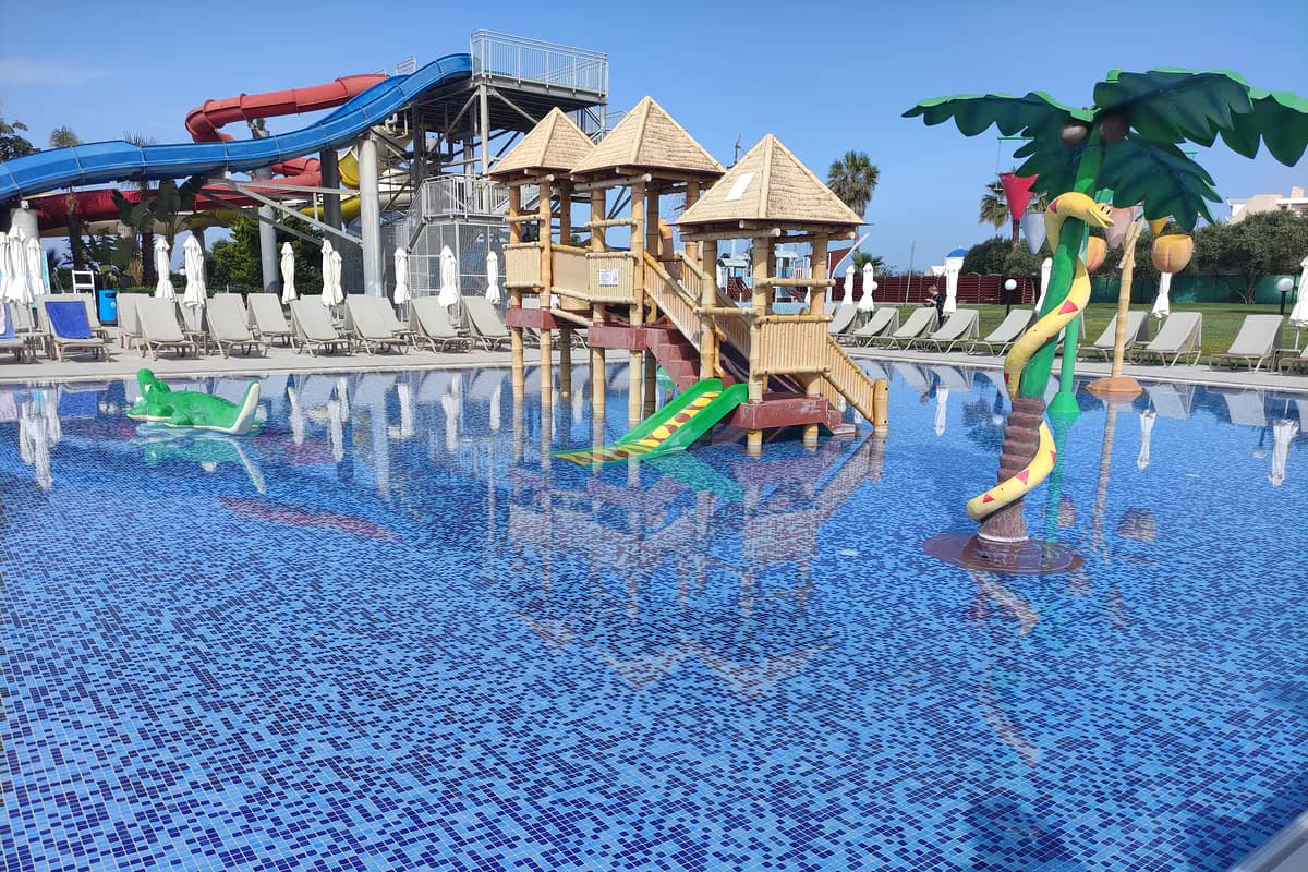 Brodzik w hotelu Louis Phaethon Beach w Pafos: drewniany domek z małymi zjeżdżalniami, figury krokodyla i węża, w tle wieża dużych zjeżdżalni