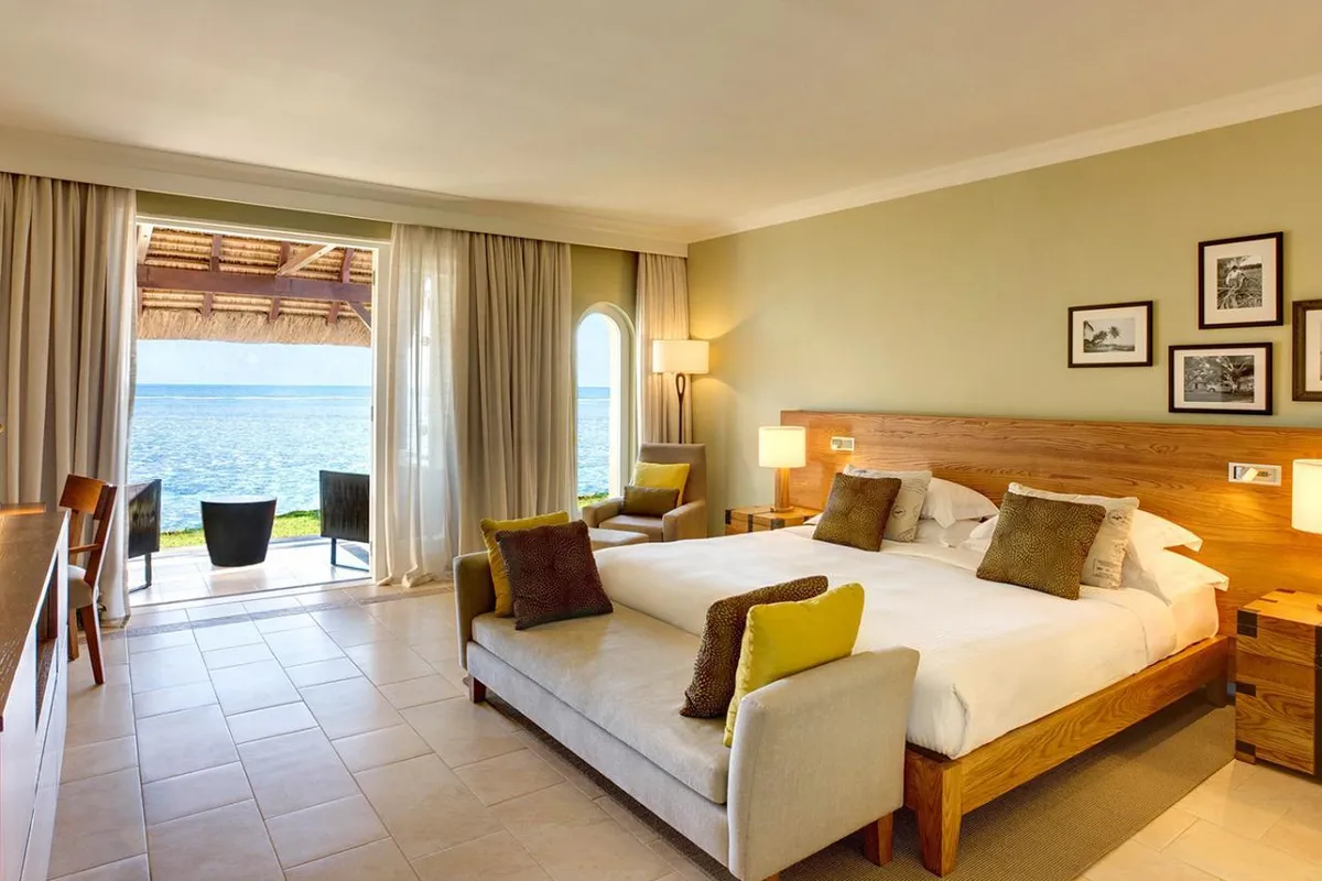 Pokój z widokiem na morze w hotelu Outrigger Mauritius Resort & Spa: szerokie łóżko, otwarte drzwi na taras z dwoma fotelami i ocean za trawnikiem