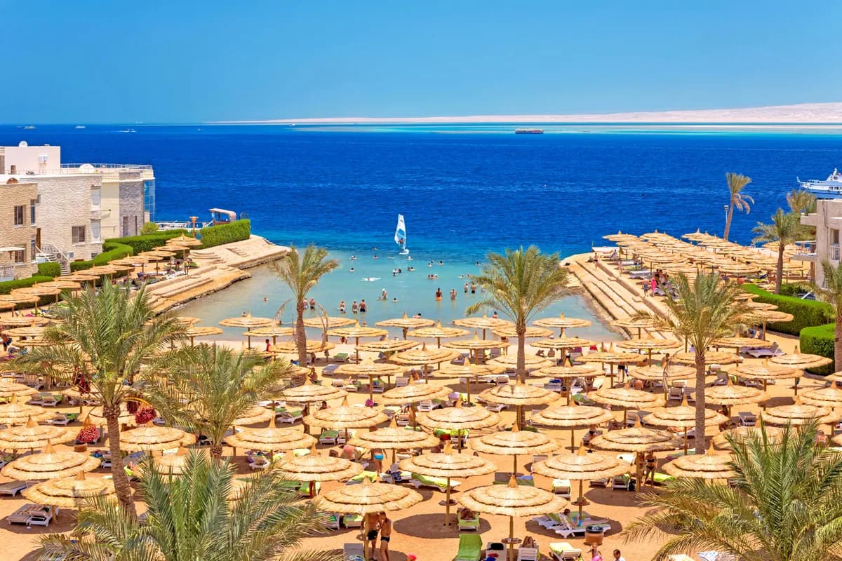 Plaża hotelu Seagull Beach Resort w Hurghadzie z parasolami z suchych liści i palmami, płytka zatoka, windsurfer i Morze Czerwone